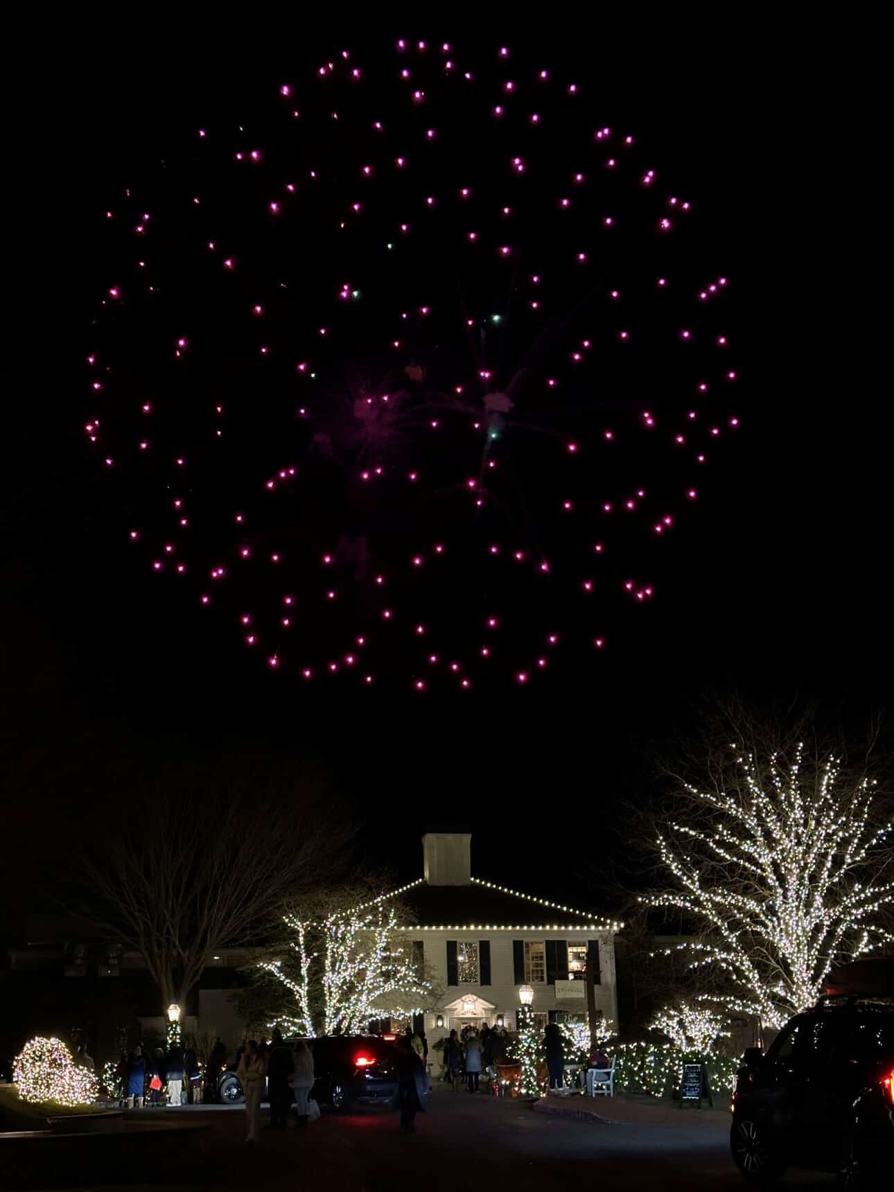 A large purple firework bursts over the Merry Market in Cape Cod, where a building is decorated with white holiday lights.