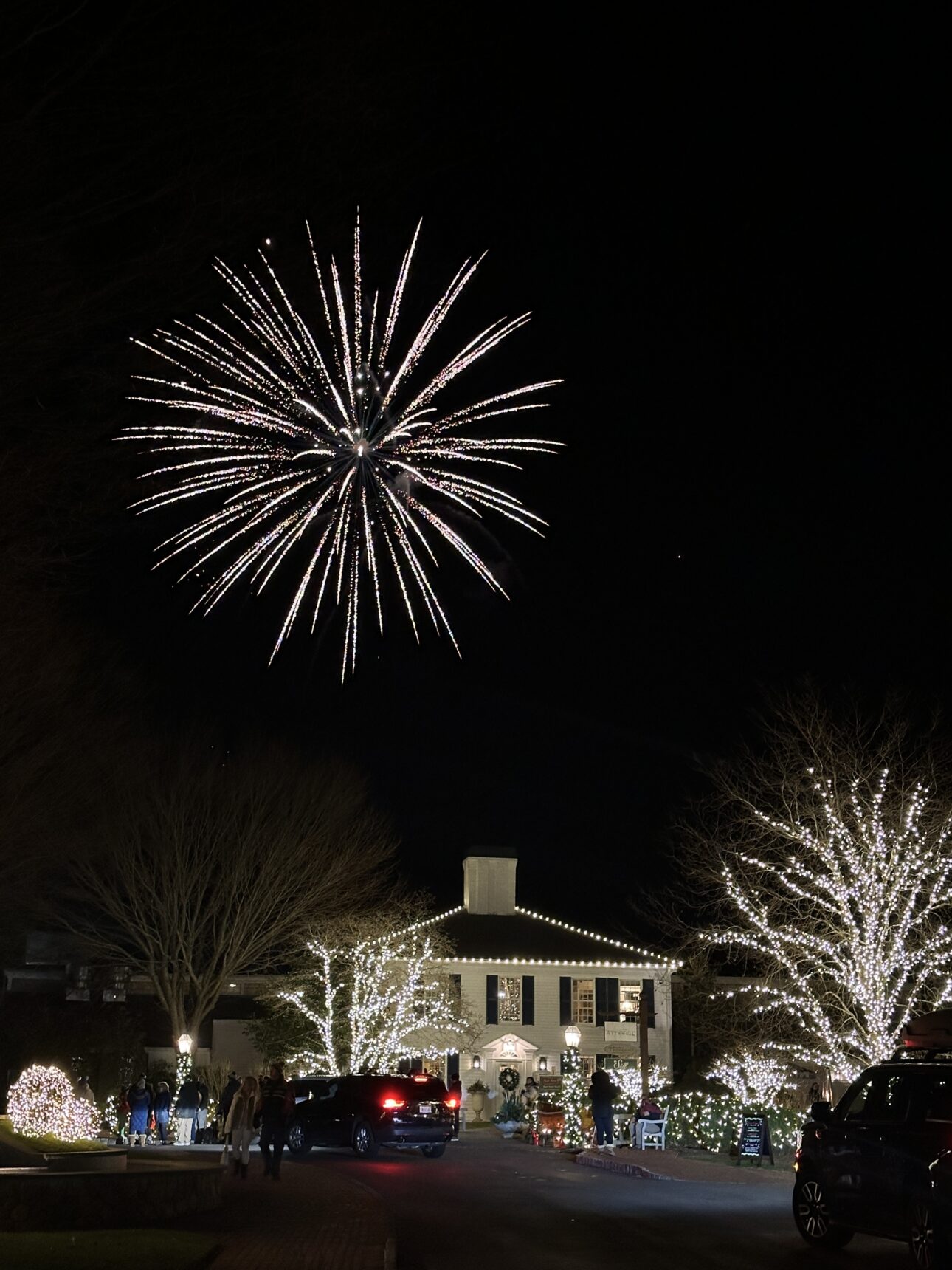 A large white firework bursts in the night sky over a Cape Cod Merry Market building decorated with Christmas lights and two illuminated trees.