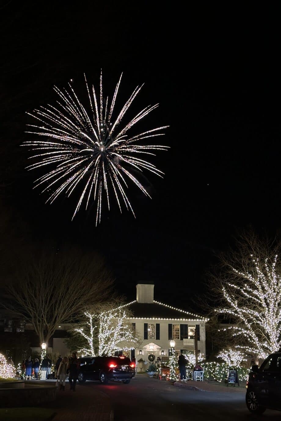 A large white firework bursts in the night sky over a Cape Cod Merry Market building decorated with Christmas lights and two illuminated trees.