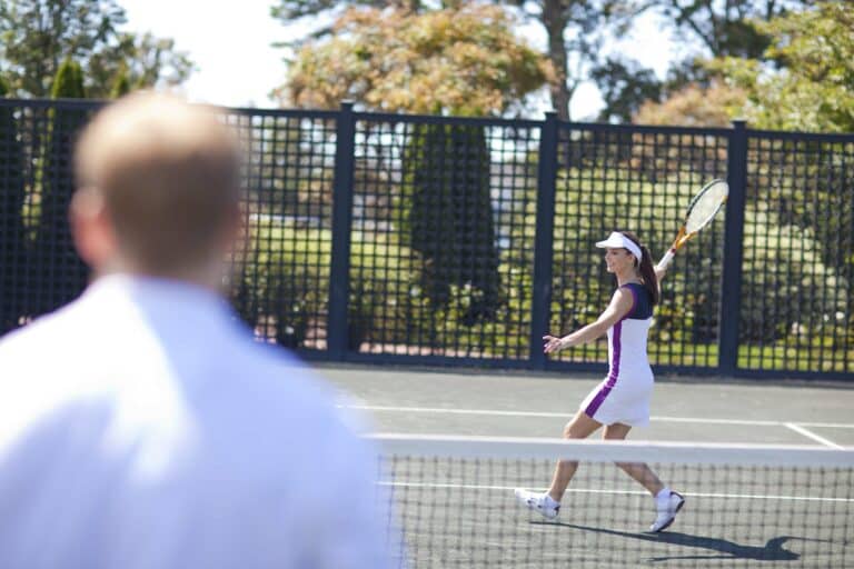 A woman in a white and purple tennis outfit prepares to hit a forehand shot during a Racquet Sports match, with a blurred opponent in the foreground.