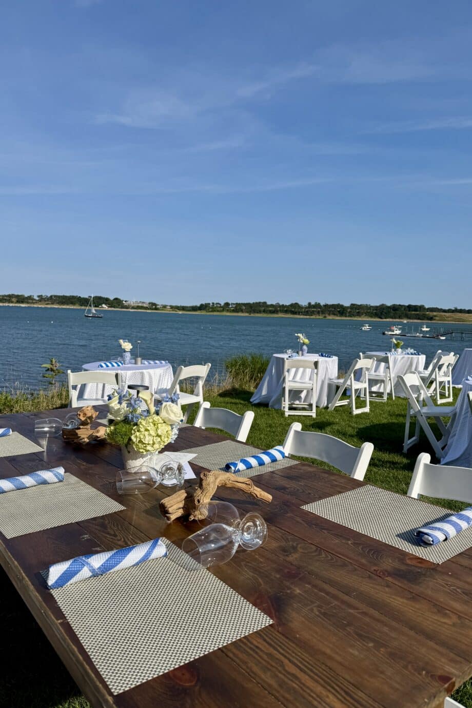 Soirée on the Bay: Outdoor event setup with wooden and white tables, chairs, placemats, striped napkins, and floral centerpieces near a lake under a clear blue sky.