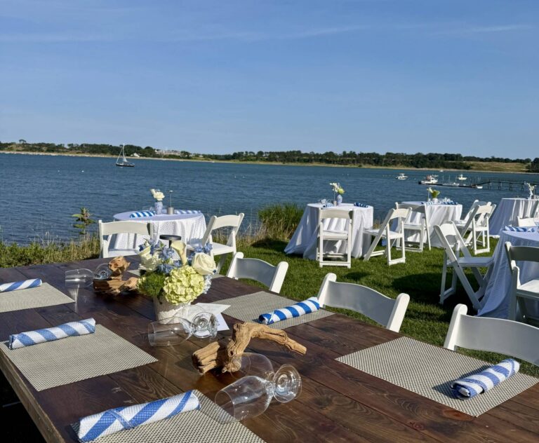 Soirée on the Bay: Outdoor event setup with wooden and white tables, chairs, placemats, striped napkins, and floral centerpieces near a lake under a clear blue sky.