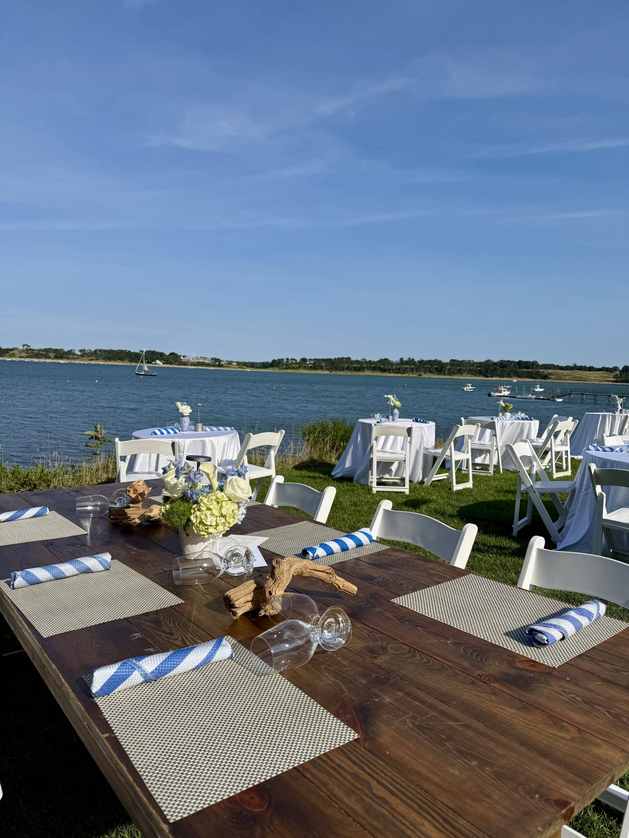 Soirée on the Bay: Outdoor event setup with wooden and white tables, chairs, placemats, striped napkins, and floral centerpieces near a lake under a clear blue sky.