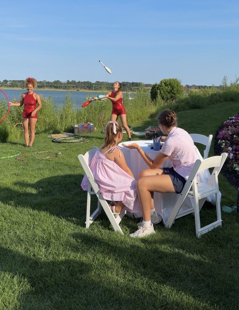 Two children sit at a table on grass while two performers in red outfits juggle and use a hula hoop beside a lake under a clear blue sky, creating a lively scene reminiscent of a Soirée on the Bay.