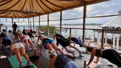A group of people practice yoga on mats under a canopy on a wooden deck overlooking a body of water and distant shore at a serene Cape Cod hotel.
