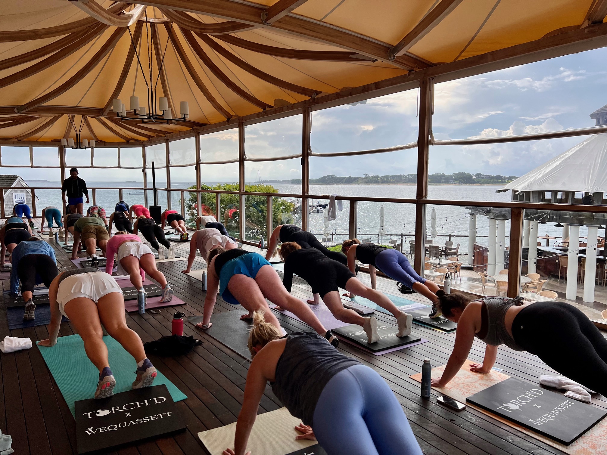 A group of people practice yoga on mats under a canopy on a wooden deck overlooking a body of water and distant shore at a serene Cape Cod hotel.