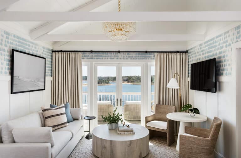 Bright living room with neutral furniture, round coffee table, and chandelier. Large windows open to a balcony with views of water and greenery—reminiscent of the serene ambiance at Wequassett Resort and Golf Club, a premier Cape Cod wedding venue.