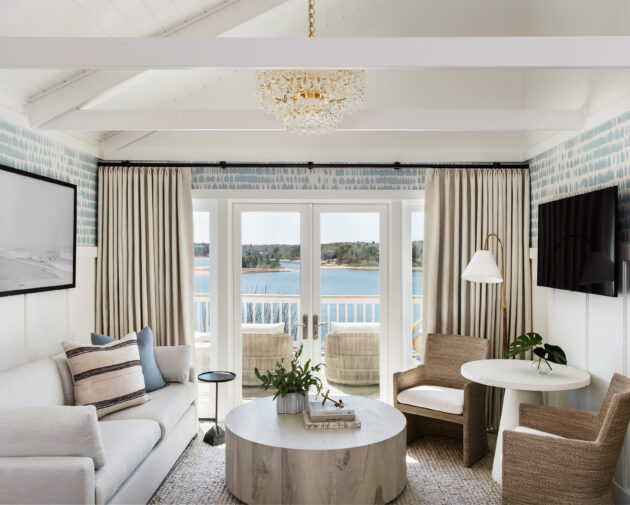 A brightly lit room in the Hydrangea Cottage with white walls, a light-colored sofa, chairs, a round coffee table, a large wall-mounted TV, and a window with curtains overlooking a body of water.