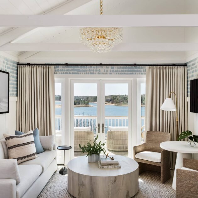 Bright living room with neutral furniture, round coffee table, and chandelier. Large windows open to a balcony with views of water and greenery—reminiscent of the serene ambiance at Wequassett Resort and Golf Club, a premier Cape Cod wedding venue.