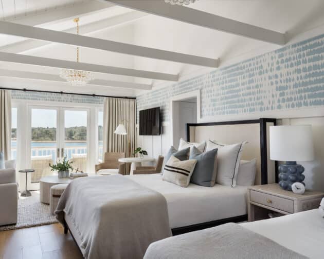 Bright hotel room with two double beds, light-colored bedding, patterned wallpaper, and a seating area. Large windows open to a balcony with views of water and greenery at the acclaimed Wequassett Resort and Golf Club.