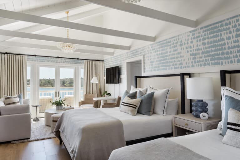 Bright hotel room with two double beds, light-colored bedding, patterned wallpaper, and a seating area. Large windows open to a balcony with views of water and greenery at the acclaimed Wequassett Resort and Golf Club.