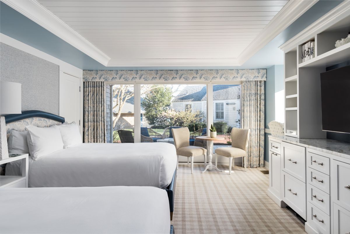 Bright hotel room with two beds, a TV, dresser, and a small table with chairs next to large windows overlooking a patio and garden.