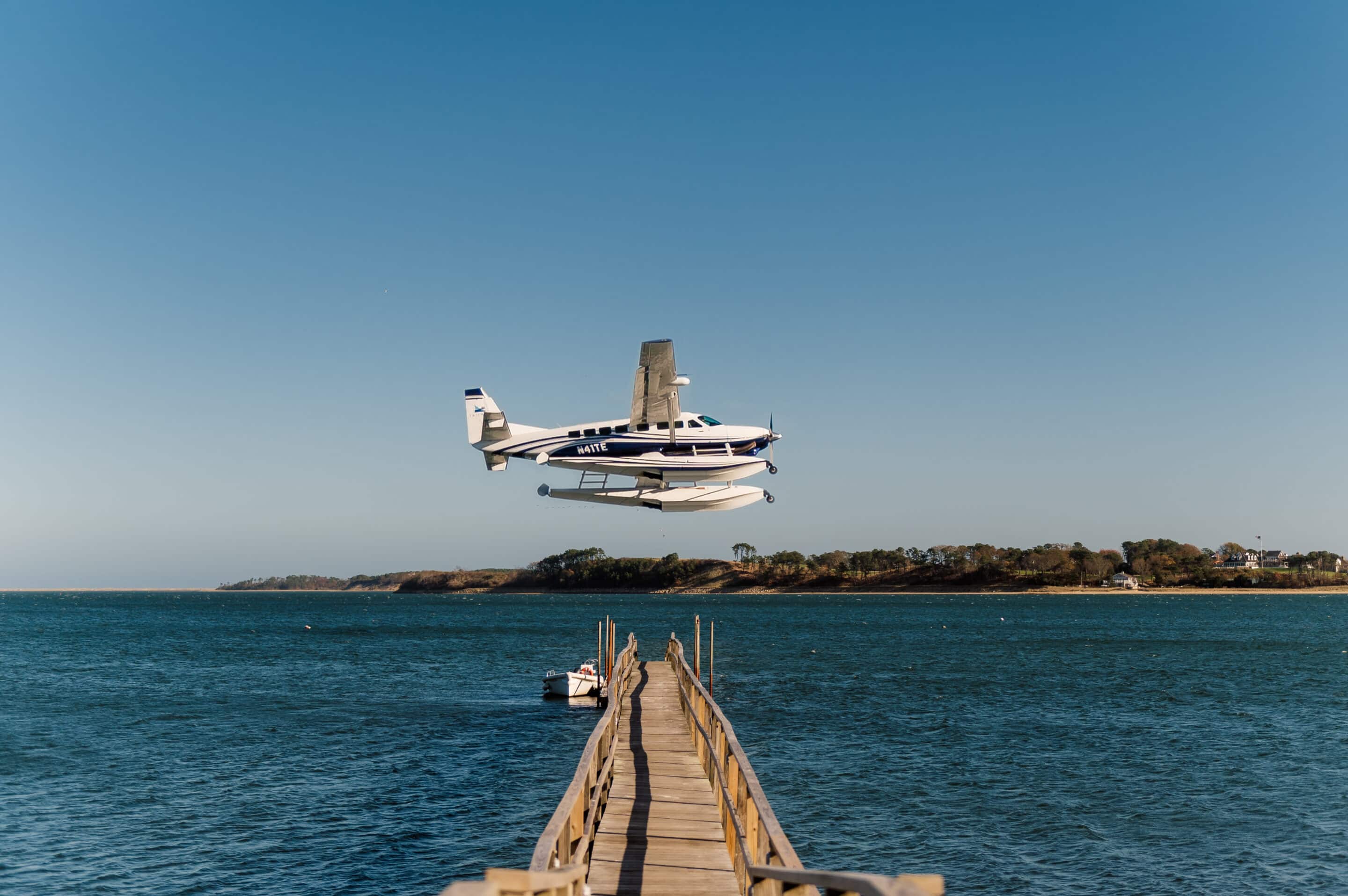 A seaplane is flying low above the water near a wooden dock extending out from Wequassett Resort into a calm bay, with land visible in the background under a clear sky.