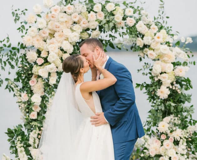 A bride and groom kiss in front of a floral arch during their outdoor wedding ceremony.