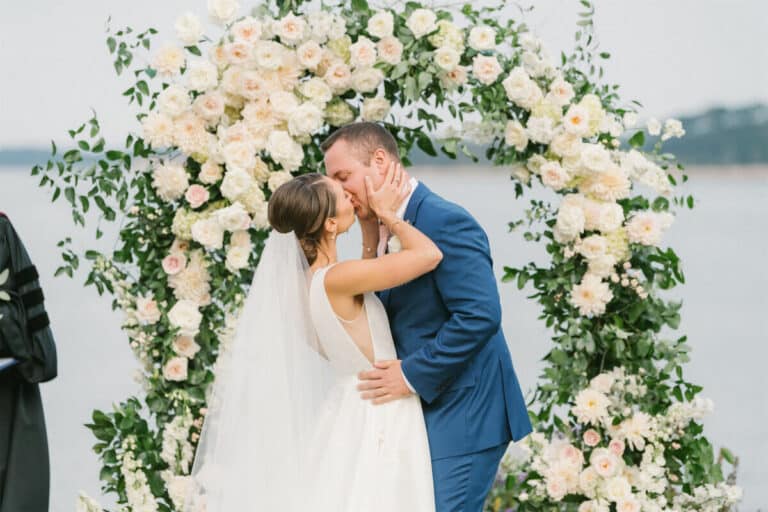 A bride and groom kiss in front of a floral arch during their outdoor wedding ceremony.