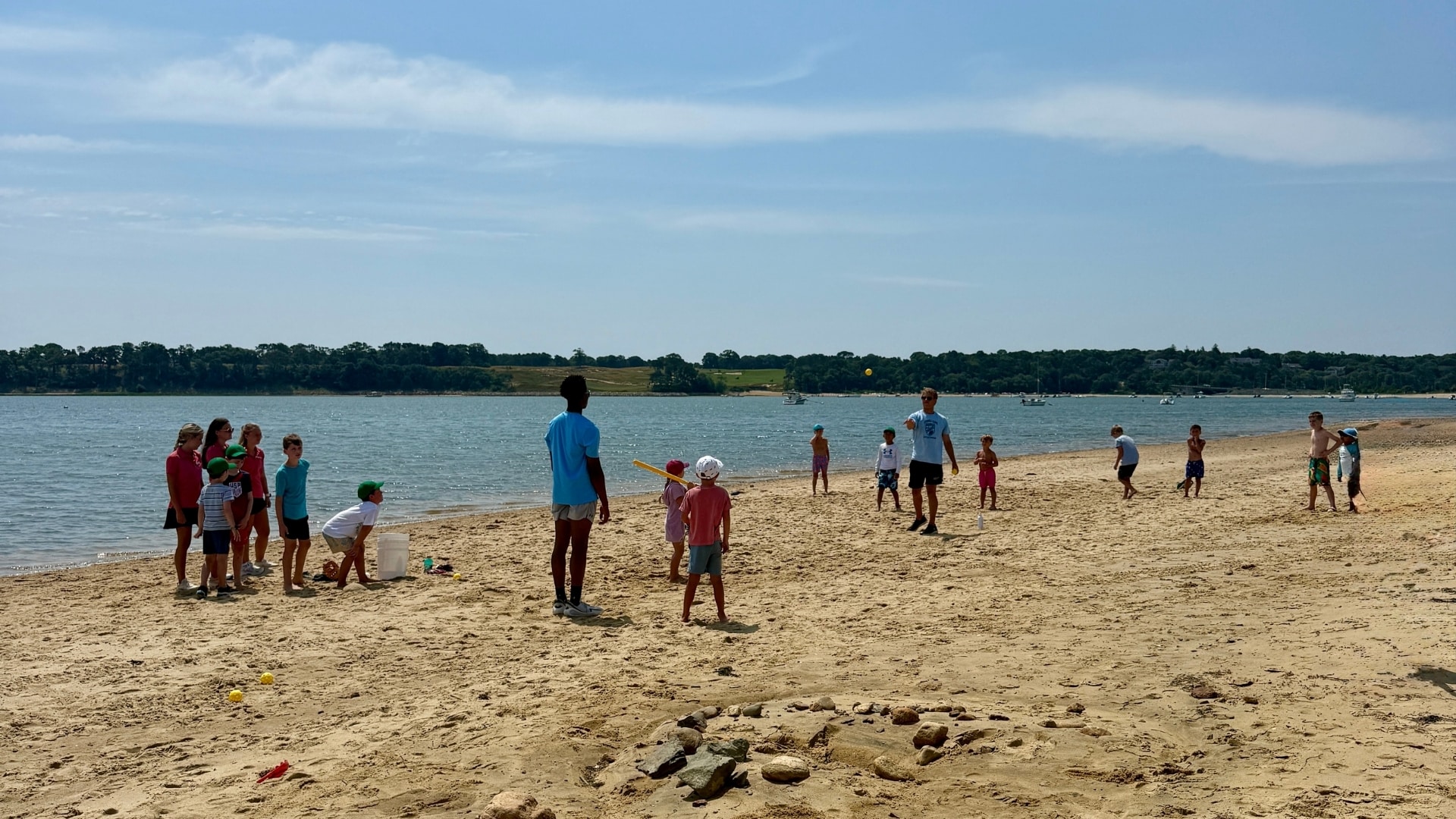 Children and adults play games and interact on a sandy beach by the water under a sunny sky, with trees visible across the water—creating a scene reminiscent of the fun and excitement found at the Wequassett Adventure Club.