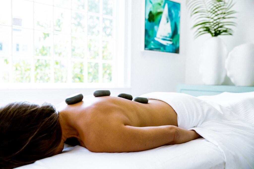 Person lying face down on a massage table with hot stones on their back, covered by a white towel, enjoying the La Bella Experience in a bright room with a painting and plant in the background.