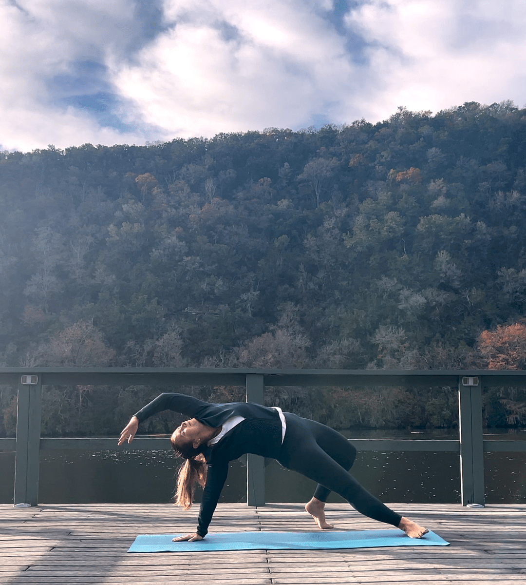 Person practicing yoga in a side plank position on a blue mat on a wooden deck at a tranquil Wellness Retreat, with a forested hillside and partly cloudy sky in the background.