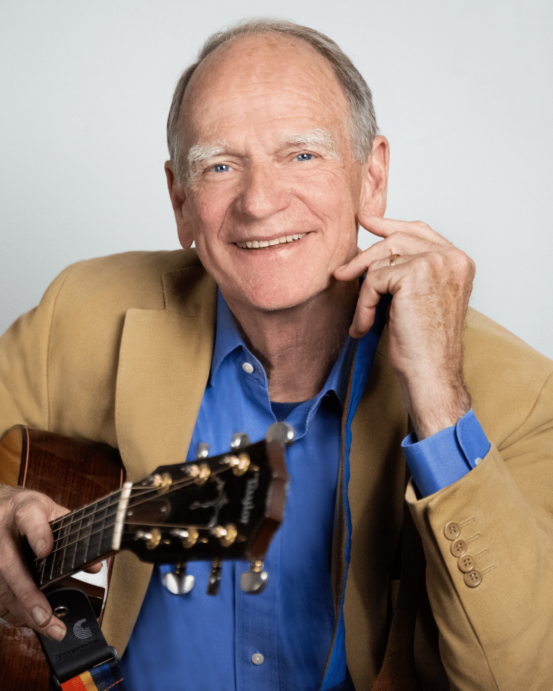 An older man in a tan blazer and blue shirt smiles at the camera while holding an acoustic guitar at the Cape Cod Jazz Festival.