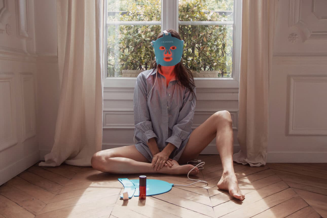A person sits on a wooden floor by a window wearing a glowing LED face mask, with skincare products and mask accessories placed in front of them.