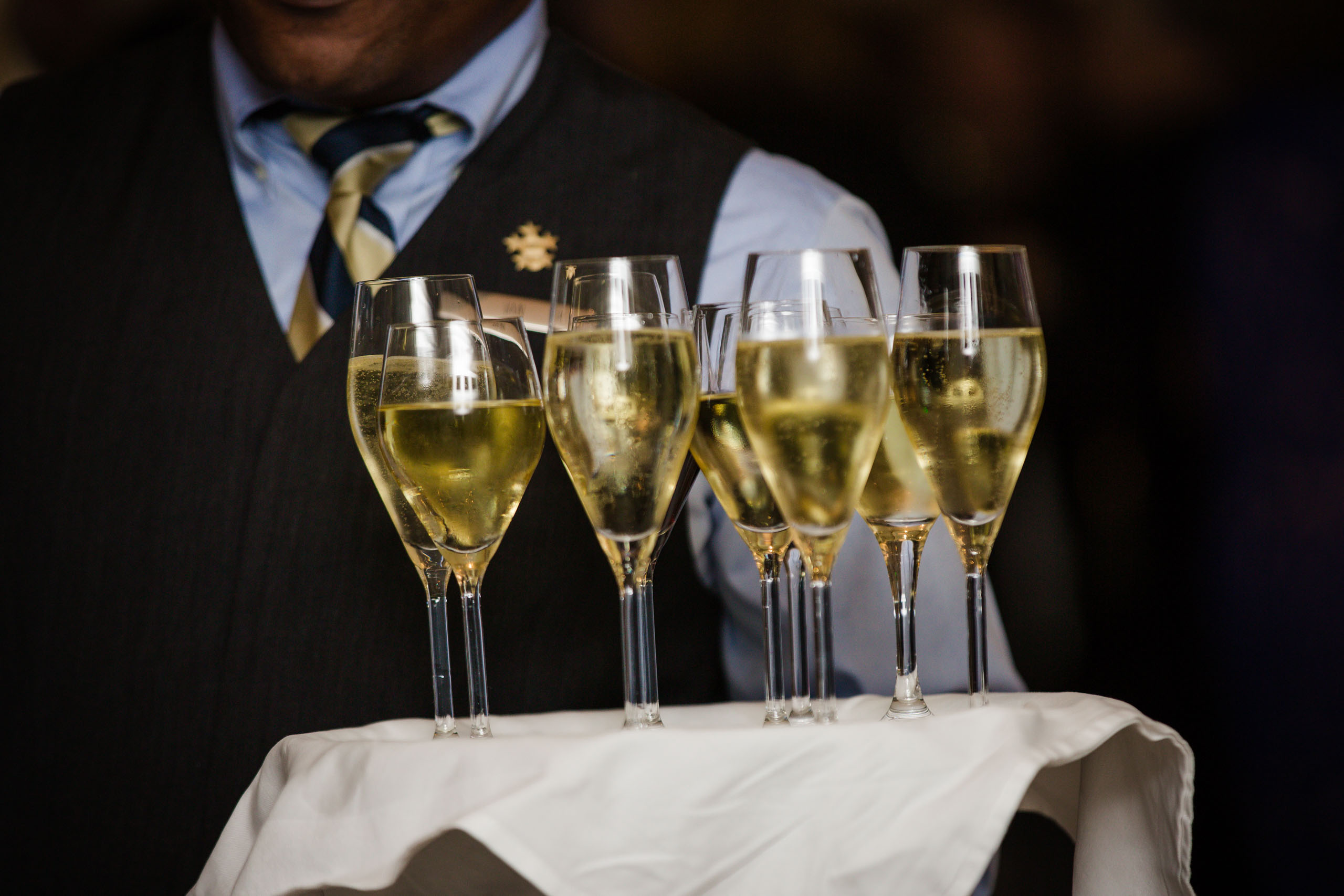 A server in formal attire carries a tray with six filled glasses of sparkling wine, covered with a white cloth.