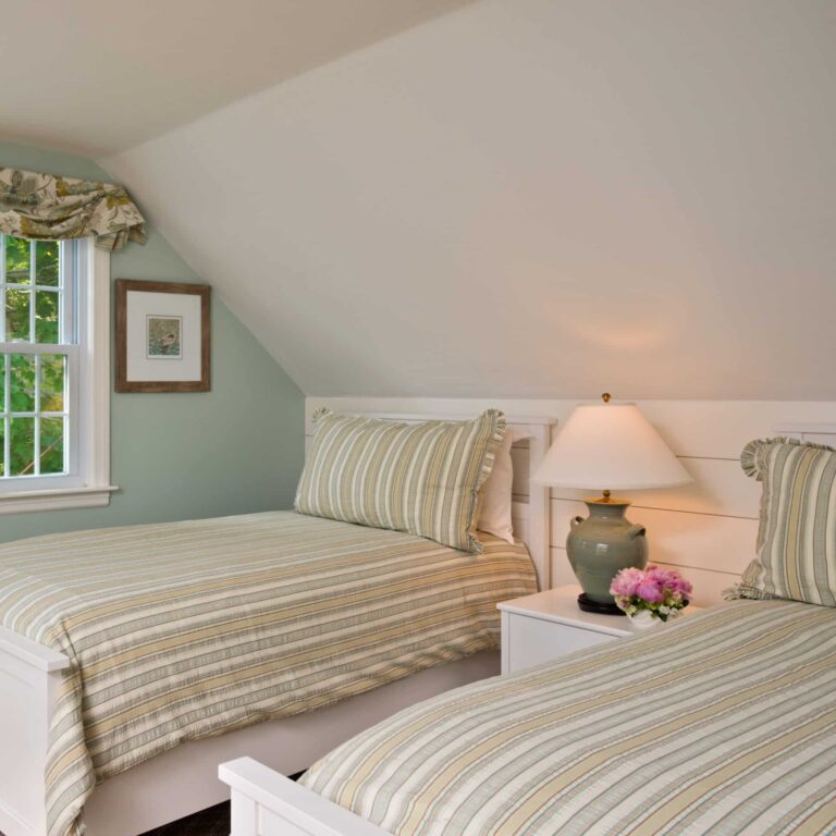 Two twin beds with striped bedding sit side by side in a cozy bedroom at a Cape Cod resort, featuring a sloped ceiling, window, nightstand, lamp, and a vase of pink flowers—perfect after a day on the Cape Cod golf course or dining at 28 Atlantic.