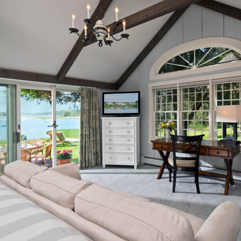 A bright living room with a beige sofa, a desk with a lamp, a TV on a white dresser, and large windows overlooking a lake and outdoor seating area offers the inviting charm of a Cape Cod resort.