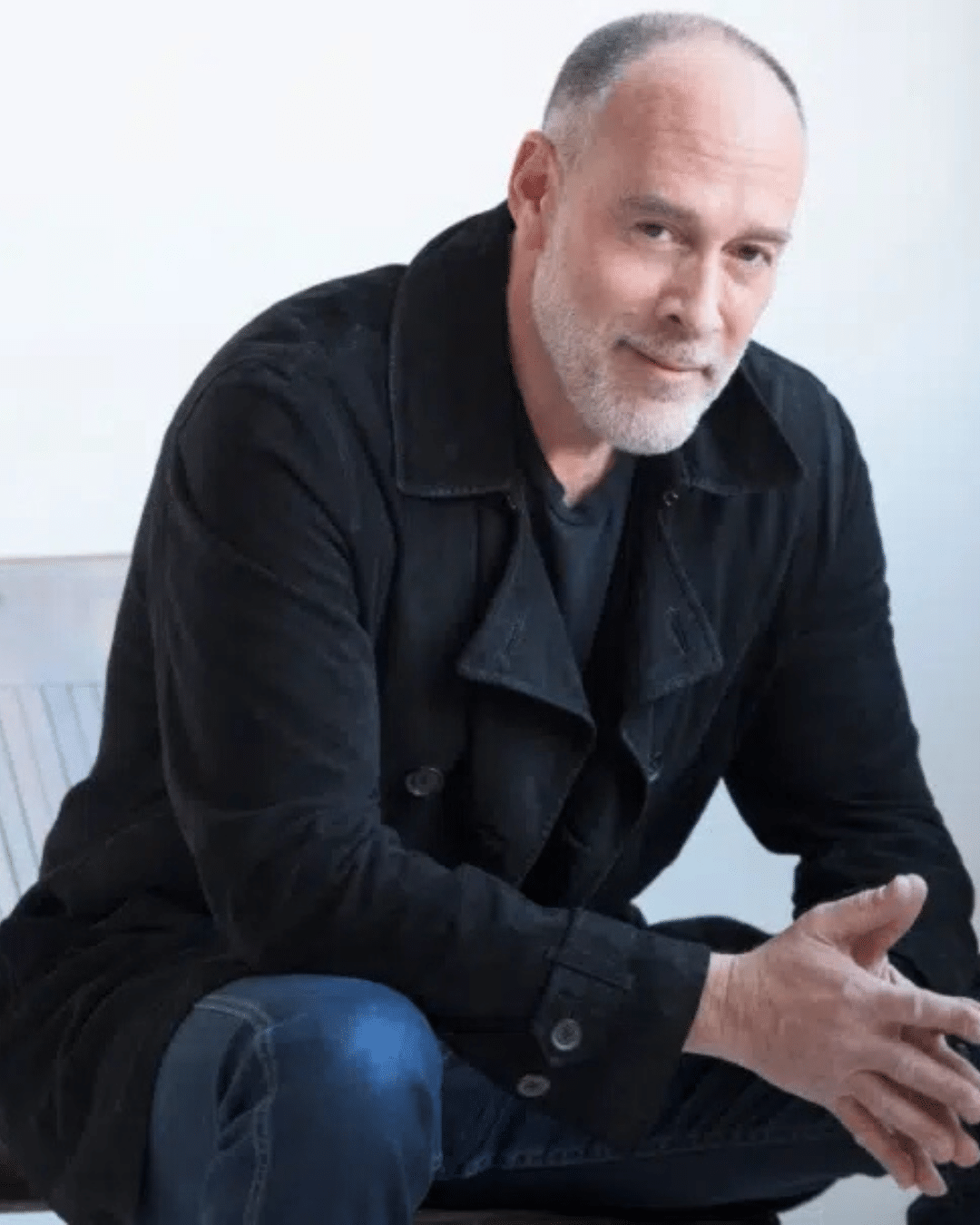 A middle-aged man with a gray beard and receding hairline wearing a black coat sits on a chair, leaning forward with hands clasped, looking at the camera, as if reflecting before his performance at the Cape Cod Jazz Festival.