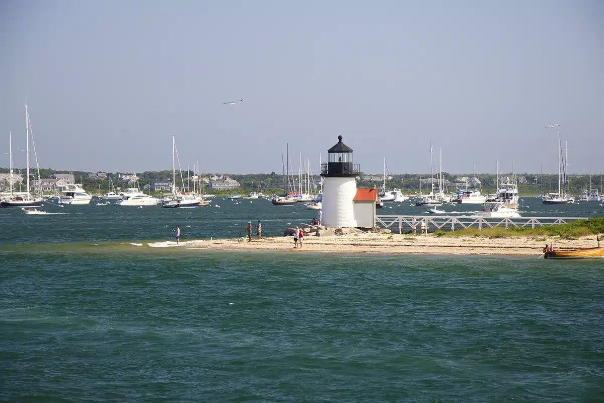 A small lighthouse stands on a narrow strip of land by the water, offering unique On the Water Experiences, with people nearby and numerous sailboats anchored in the harbor in the background.