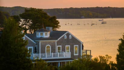 A gray house with white trim overlooks a calm bay with anchored boats at sunset on Cape Cod, surrounded by trees and hills—an inviting setting for intimate meetings or memorable events.