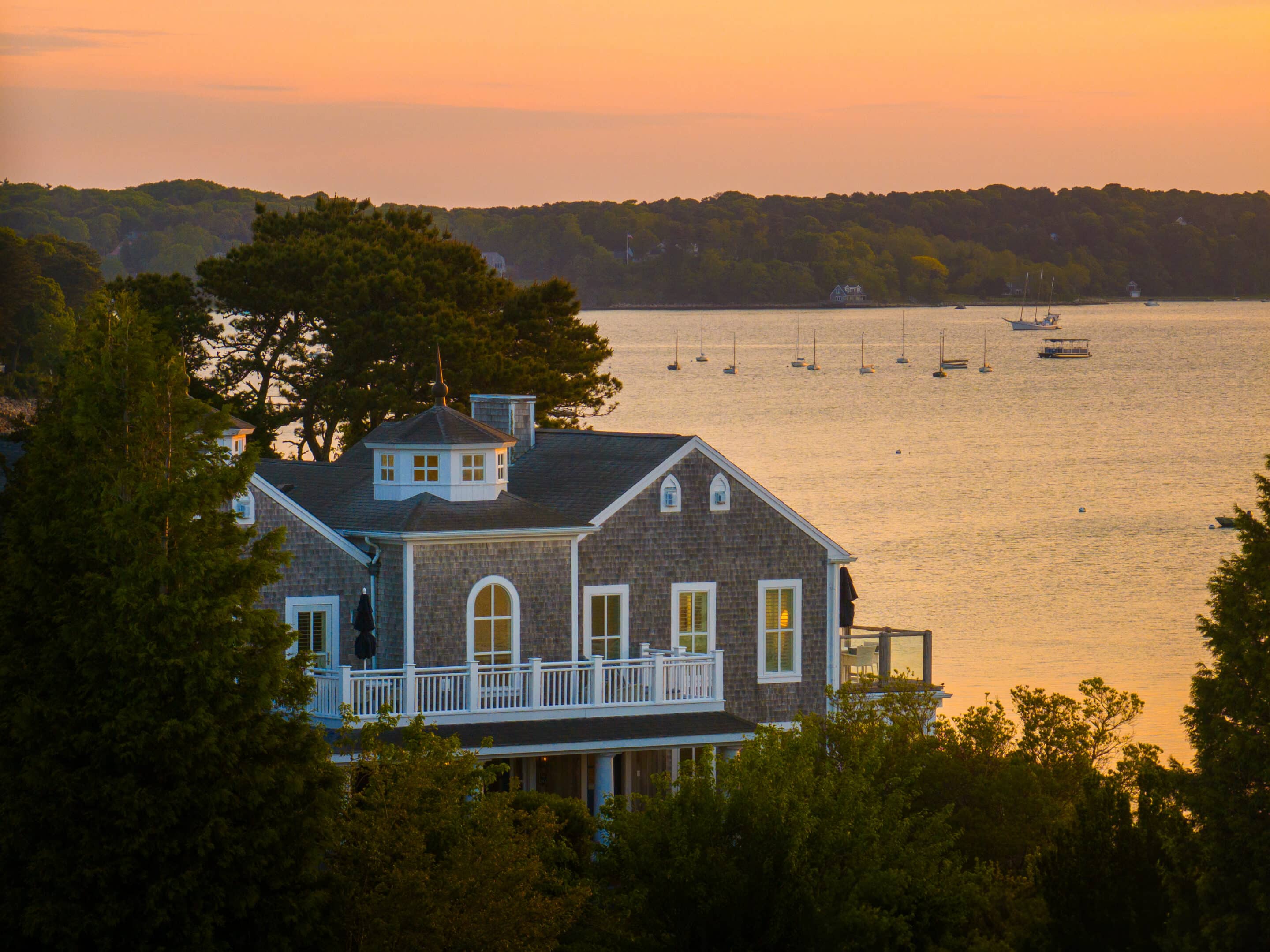 A gray house with white trim overlooks a calm bay with anchored boats at sunset on Cape Cod, surrounded by trees and hills—an inviting setting for intimate meetings or memorable events.