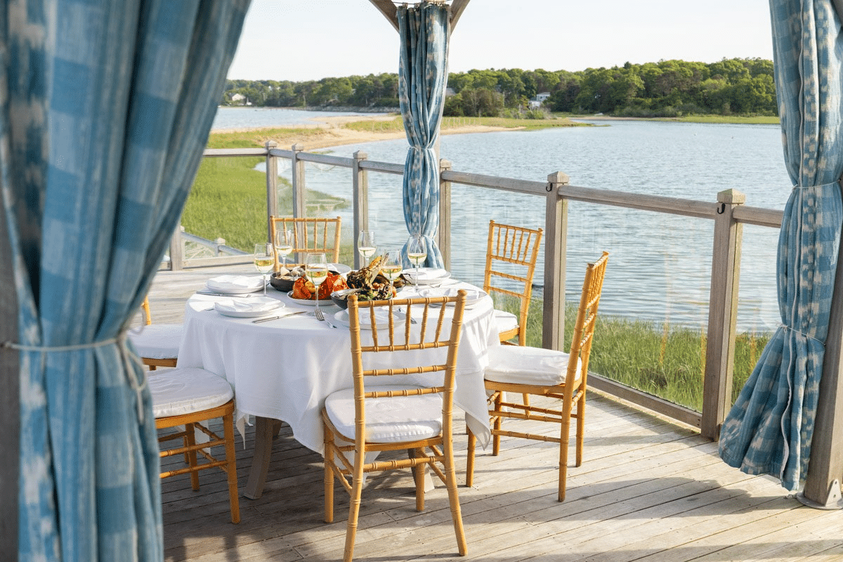 Round table with chairs, set for a meal with seafood and wine—perfect for a clambake—on a wooden deck overlooking water, with blue curtains framing the view in your own private cabana.