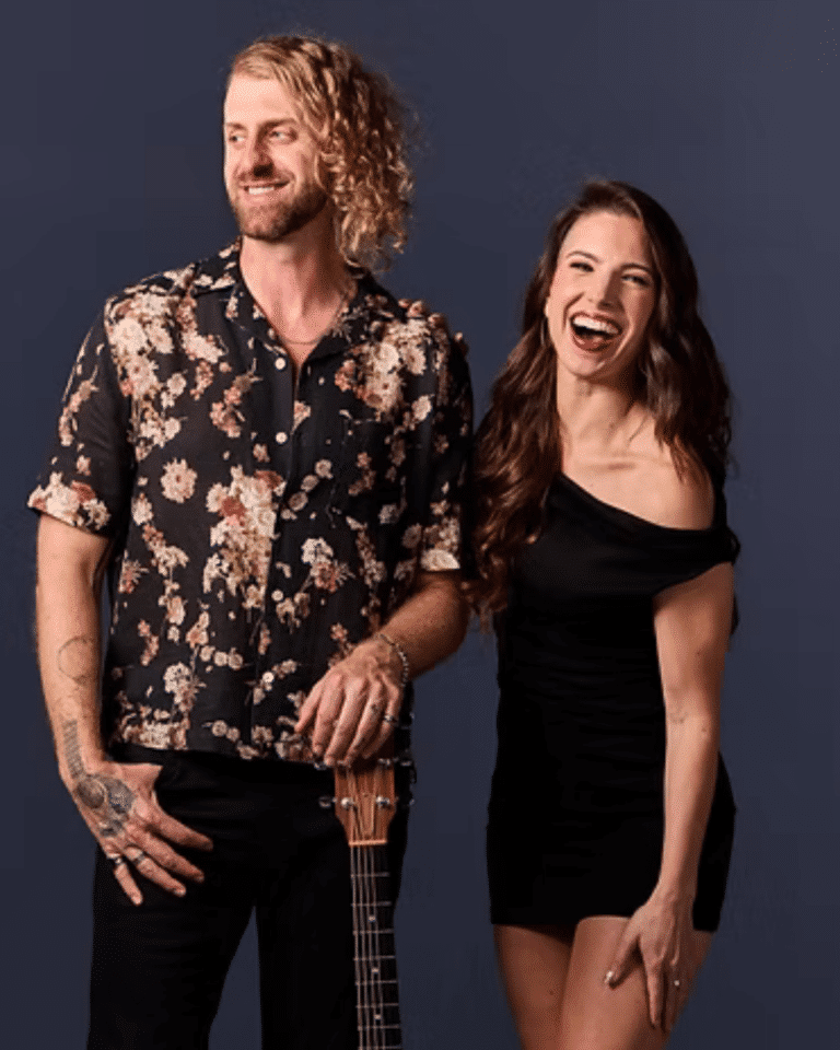A man in a floral shirt stands holding a guitar next to a woman in a black dress, both smiling against a plain dark background—ready to bring some Jazz flair to the Cape Cod Arts Festival.