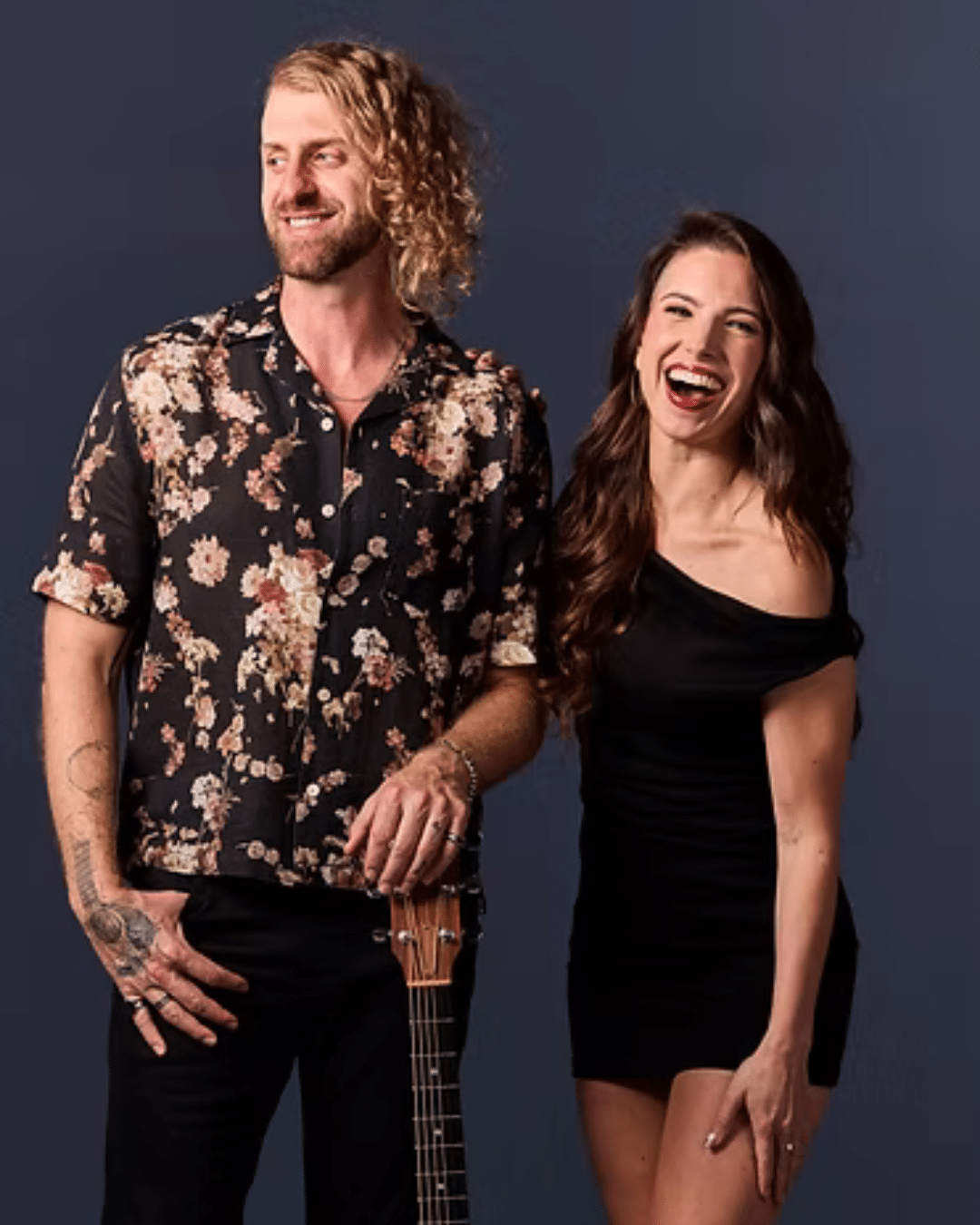A man in a floral shirt stands holding a guitar next to a woman in a black dress, both smiling against a plain dark background—ready to bring some Jazz flair to the Cape Cod Arts Festival.