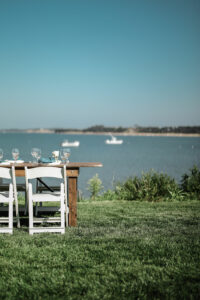 A wooden table set with glassware and white chairs sits on grass, overlooking a calm body of water at 28 Atlantic, the perfect Cape Cod wedding venue with boats drifting in the distance under a clear blue sky.