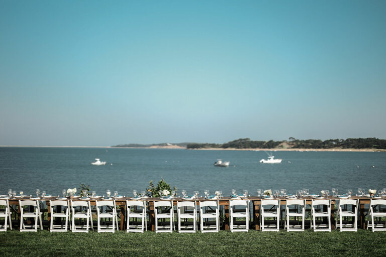A long dining table with white chairs is set up outdoors on grass, overlooking calm water and boats—an idyllic scene perfect for a Cape Cod wedding venue or an event at Wequassett Resort and Golf Club.