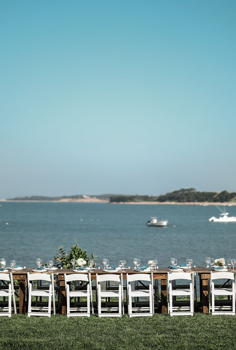 A long dining table with white chairs is set up outdoors on grass, overlooking calm water and boats—an idyllic scene perfect for a Cape Cod wedding venue or an event at Wequassett Resort and Golf Club.