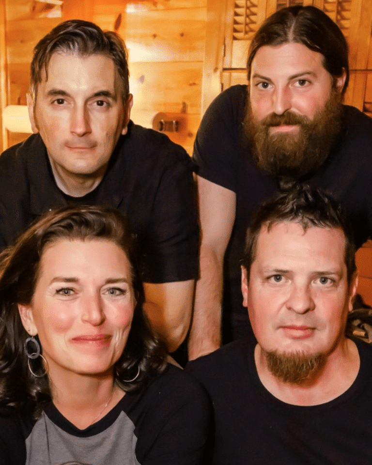 Four adults, three men and one woman, pose closely together indoors in warm lighting at a Cape Cod Arts Festival, all facing the camera and smiling slightly.