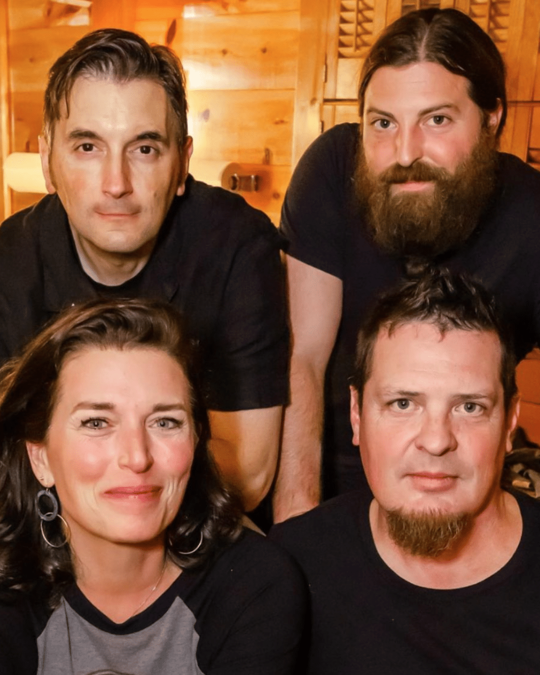 Four adults, three men and one woman, pose closely together indoors in warm lighting at a Cape Cod Arts Festival, all facing the camera and smiling slightly.