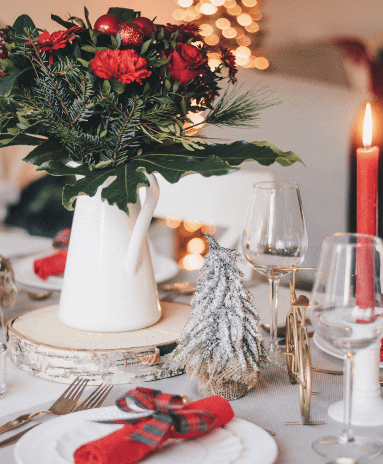A festive table setting features a white pitcher with red flowers, a small frosted tree, red candles, wine glasses, and red plaid napkins—perfect for gathering after the town’s Tree Lighting, with a blurred Christmas tree in the background.
