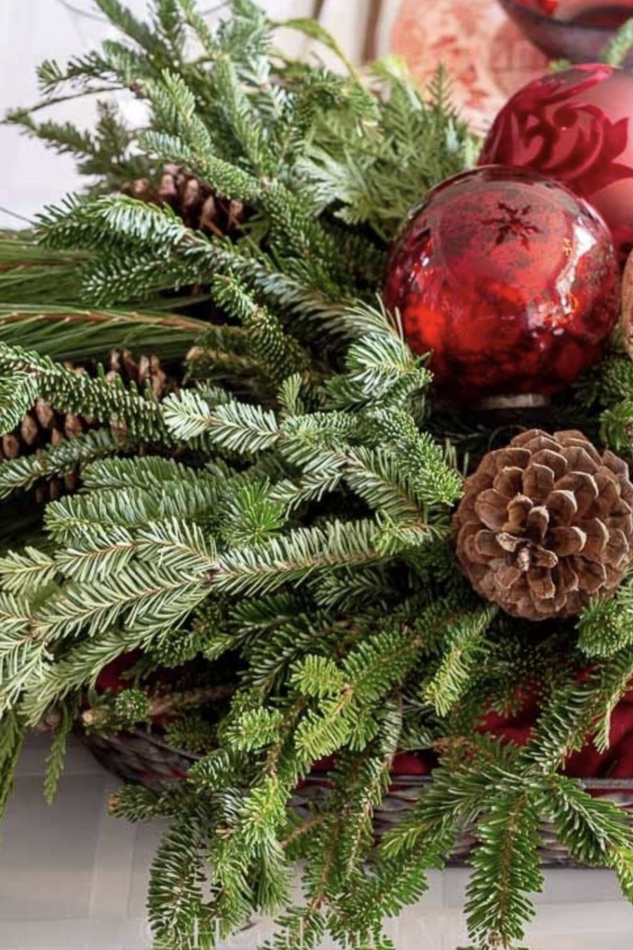 A festive arrangement reminiscent of Mrs. Claus’s cottage, featuring pine branches, pine cones, and red and gold Christmas ornaments in a basket on a white surface.