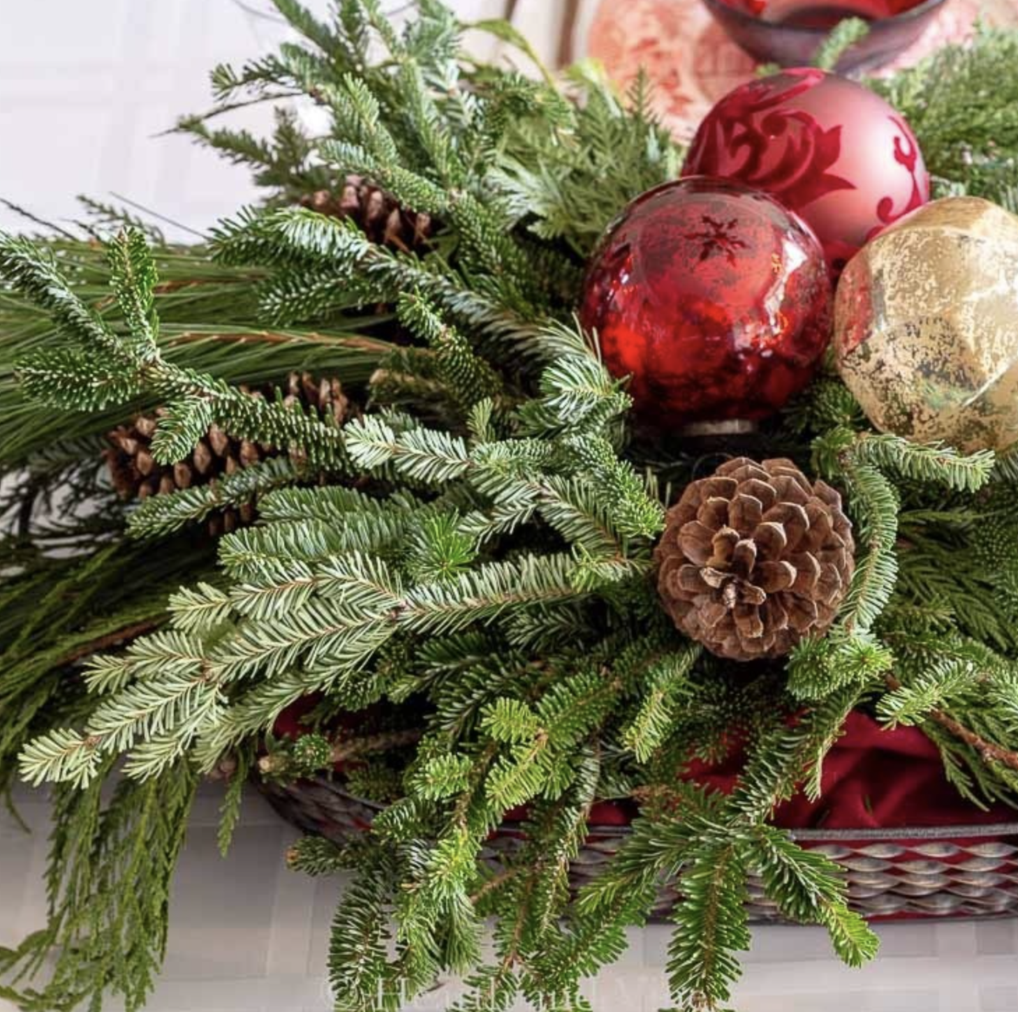 A festive arrangement reminiscent of Mrs. Claus’s cottage, featuring pine branches, pine cones, and red and gold Christmas ornaments in a basket on a white surface.