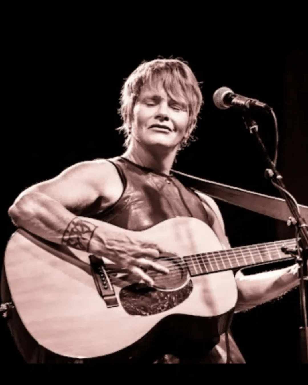 Person playing an acoustic guitar and singing into a microphone on stage at the Cape Cod Jazz Festival, eyes closed and wearing an expressive facial expression.