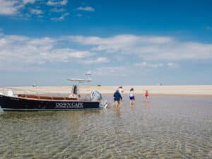 A small group of people begins their journey from a beached boat labeled "Down Cape Charters & Boat Rentals," walking across shallow water toward the sandy Outer Beach under a clear blue sky.