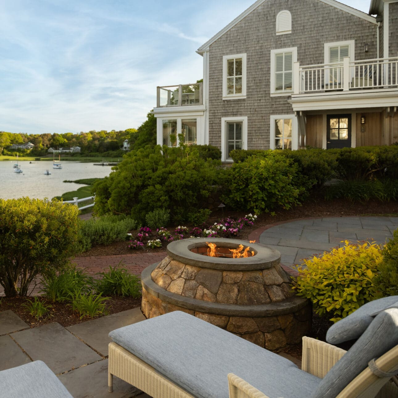 Stone fire pit with flame on a patio, two lounge chairs in the foreground, gray shingle house and waterfront with boats in the background, surrounded by greenery and flowers—an inviting outdoor space perfect for enjoying the tranquil ambiance of the Signature Water View Suite.