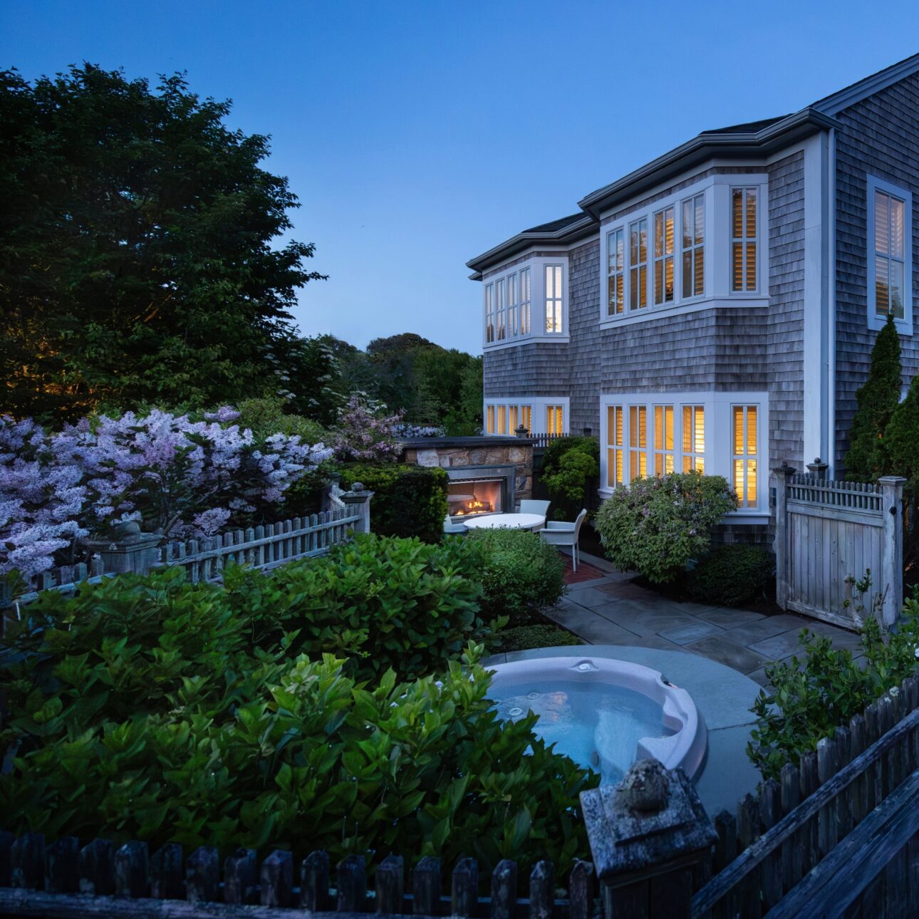 A two-story house with large windows, wooden siding, garden plants, a picket fence, an outdoor fireplace, and a round Outdoor Spa hot tub at dusk—part of the Signature Collection.