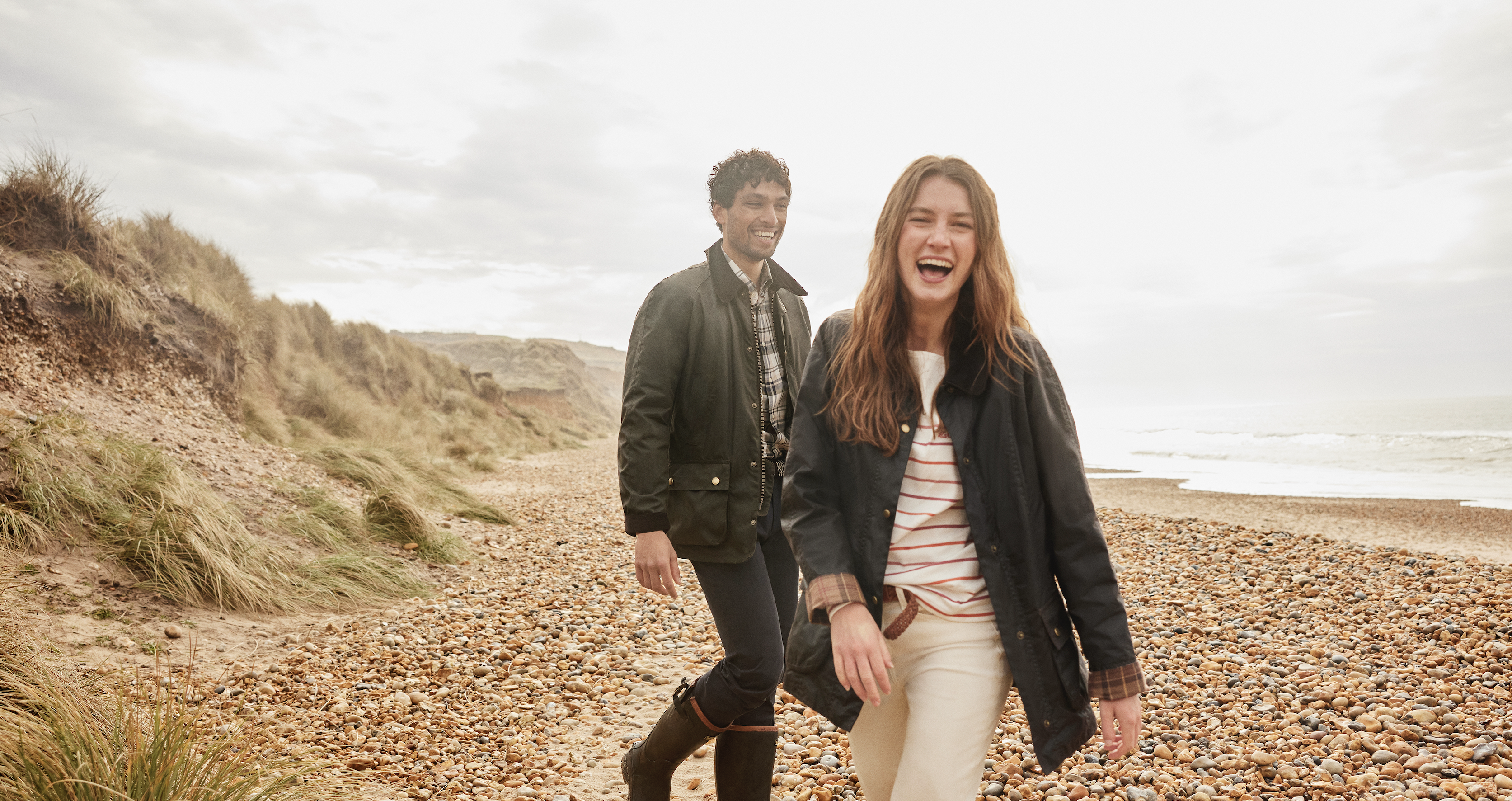 A man and a woman walk along a rocky beach, both smiling and dressed in Barbour clothing. The sky is overcast.