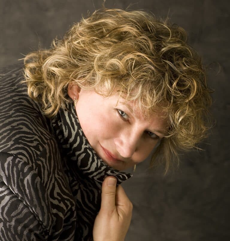 Person with short curly blond hair looks at the camera, holding up the collar of a patterned jacket, evoking the creative spirit of an Arts Festival, against a plain dark background.