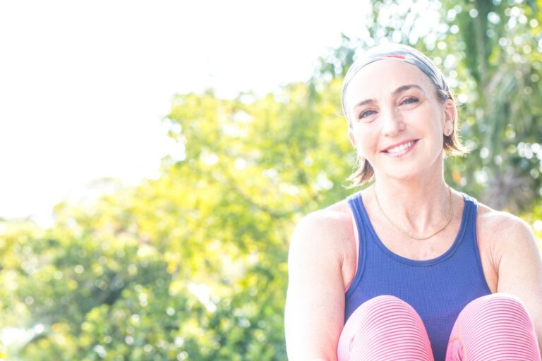 A woman wearing a headband and athletic clothes sits outdoors, smiling, with green trees and bright sunlight in the background.