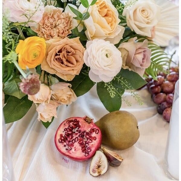 A Thanksgiving floral arrangement with roses and assorted flowers sits on a table next to grapes, a halved pomegranate, a fig, and a pear.